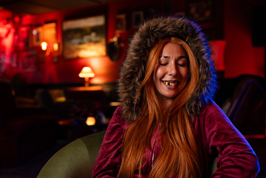 Miss Kaninna sits in an armchair in a room with red lighting and sings.