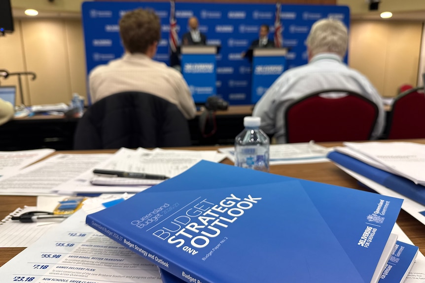 Blue books on a wooden table with a press conference blurred in the background.