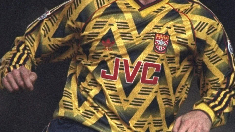 An Arsenal player wearing a black and yellow away strip, from the early 1990s, with the old red badge on it.