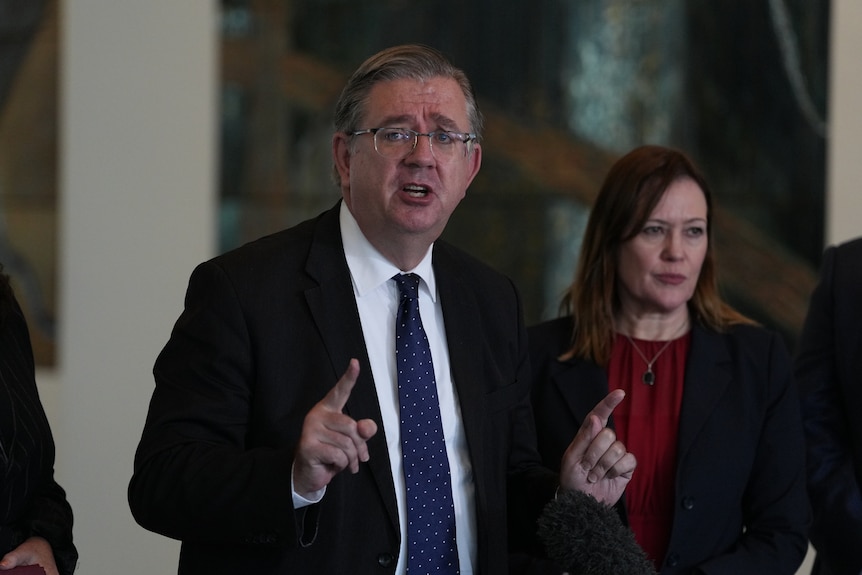A middle-aged man in a dark suit gestures as he speaks.