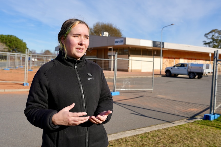 A woman in a black jumper outside fenced off buildings.