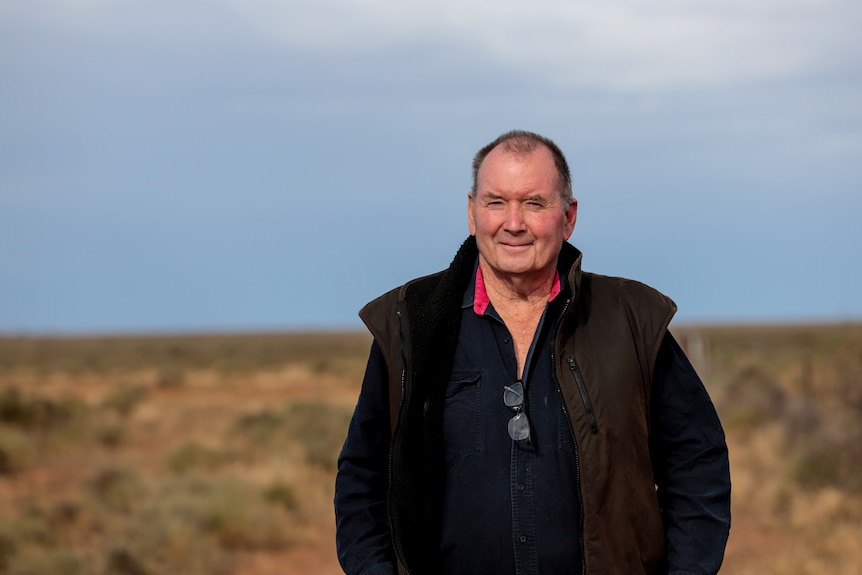 A man wearing a dark coloured shirt and vest stands in a field with the sky and horizon visible behind him.