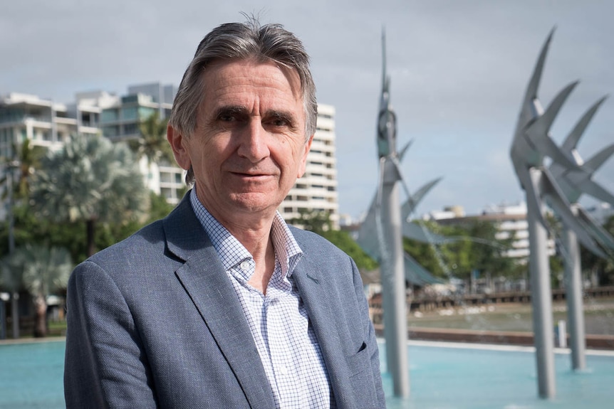 A man with grey hair wearing a grey suit stands at the Cairns lagoon with sculptures in the background.