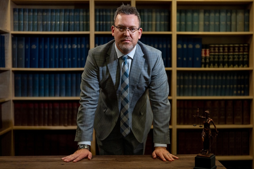 A man in a business suit stands and places both hands on a table, leaning forward, in front of bookshelves full of case law