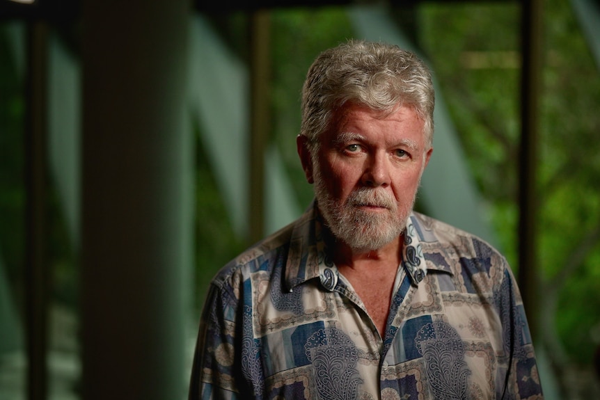 A man with white wavy hair and a white beard in a blue, biege and white patterned shirt stares in front of a green backdrop.