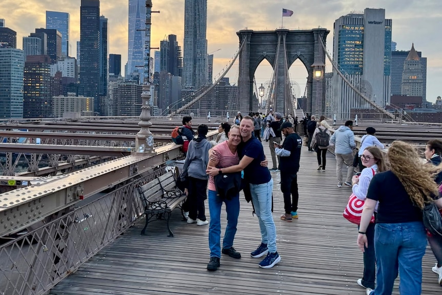 Two men embrace while standing on a bridge with the city skyline behind them