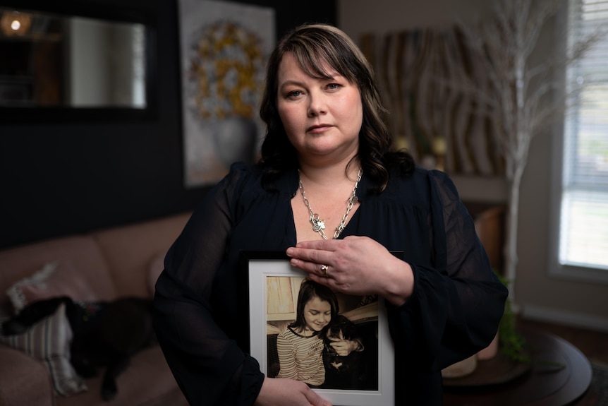 a woman holds a framed picture of a young girl looks at the camera while sitting indoors at a house