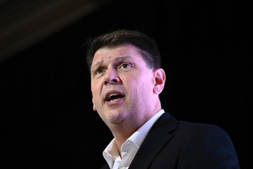 A dark-haired man in a dark suit in mid-sentence.