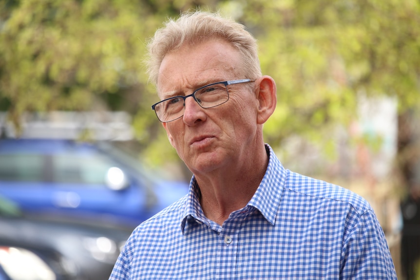 A man with short blonde hair and glasses stands outdoors looking slightly confused.