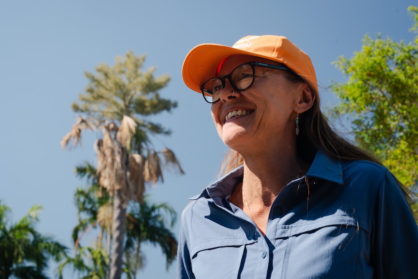 A woman wearing a cap and reading glasses, smiling as she stands outside among tall palm trees.