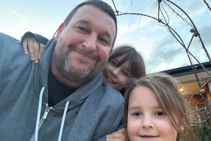 selfie of Steve with his two children, blue sky behind, a trellis with some wires.