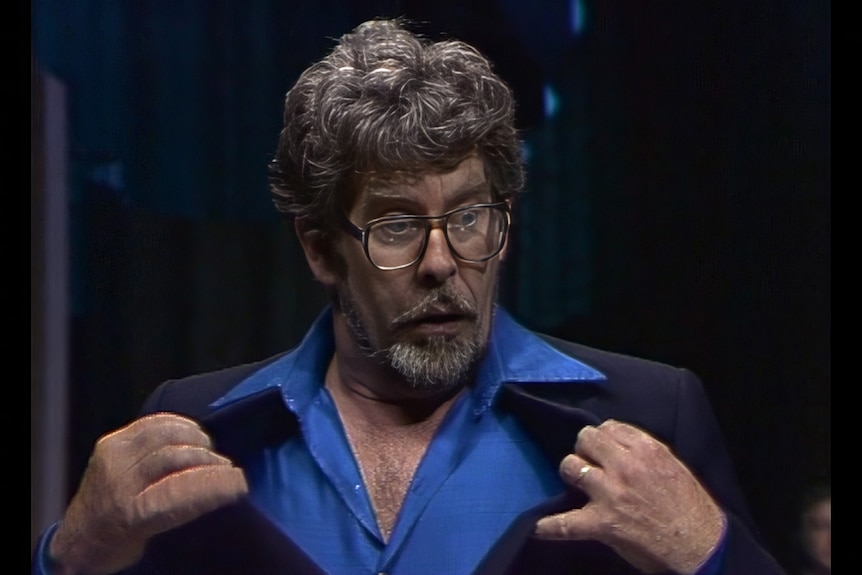 A man in a blue and black suit pulls at his collar with both hands.