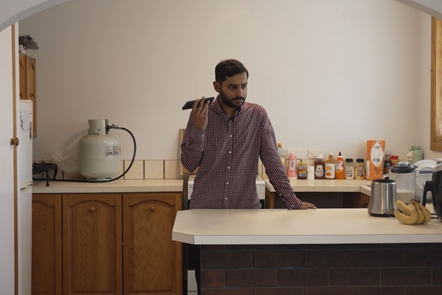 A man with combed black hair and stubble in a shirt stands in his kitchen holding his phone to listen to a voice note.