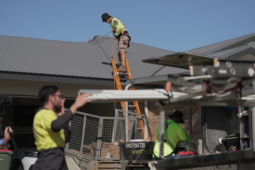 Men in yellow hi-vis shirts hold a solar panel as a man stands on a ladder at roof height next to a house.