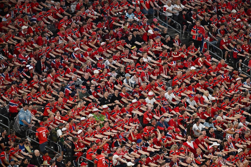 a packed crowd in a stadium all wearing red Norway jerseys miming a rowing action