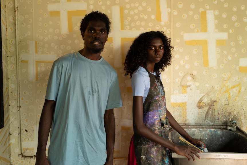 A young man wearing a t-shirt, and a woman wearing a painter's smock, standing in front of a wall painted with crosses and dots.