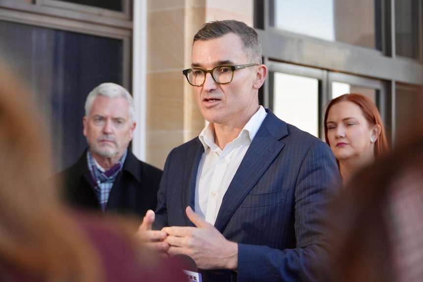 John Carey gestures with his hands while pictured at a press conference