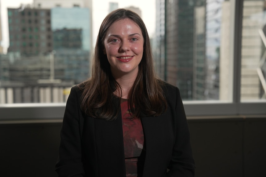 A white woman with long brown hair, black blazer and red shirt, sitting in front of window of high-rise building.