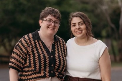 Two sisters smiling at the camera in front of a small forest.