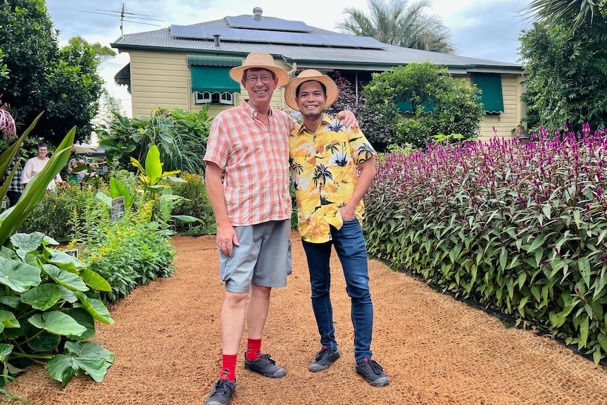 Jerry Coleby-Williams stands with his arm around partner Thuan, both smiling in a bright garden in front of a house