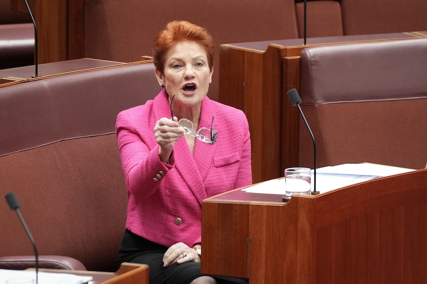 A woman in a pink blazer gestures with her glasses from a bench.