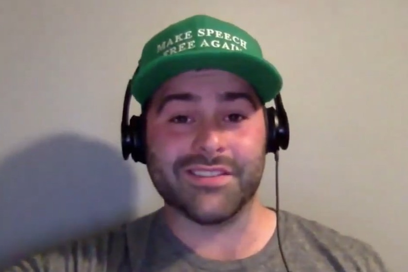 Man in grey shirt with headphones and green hat that says Make Speech Free Again