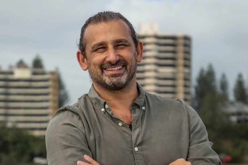 A middle aged man smiling at the camera, arms crossed, apartment buildings and houses in the background