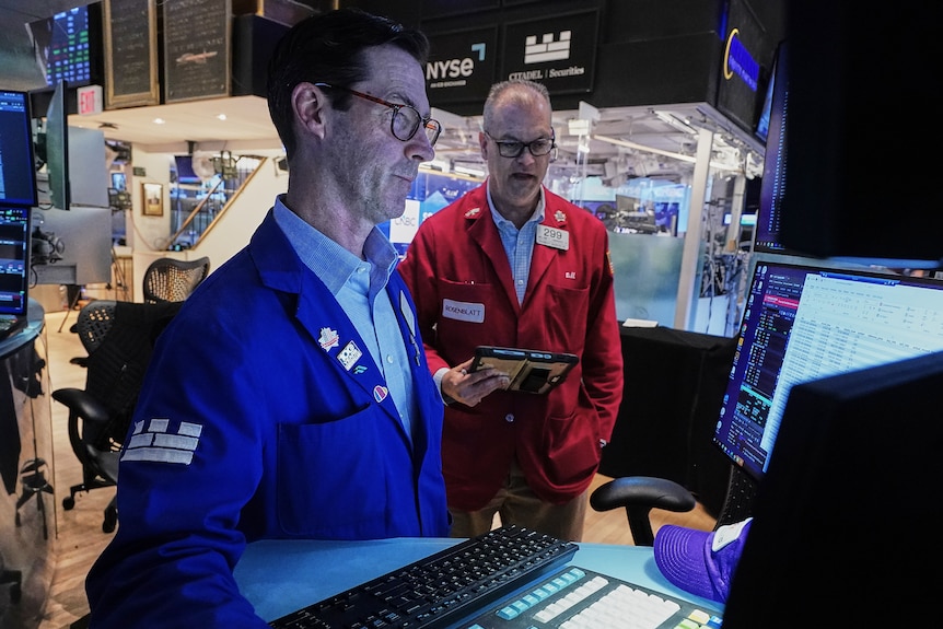 A man wearing in a blue jacket and a man in a red jacket, both wearing dark glasses, looking at illuminated computer screens.