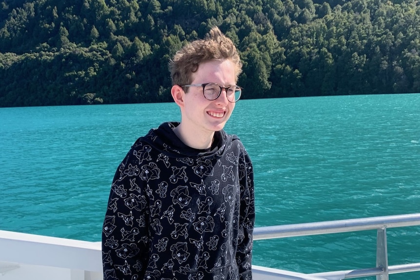A young man with glasses standing on a boat. Behind him is a large mountain and water. 