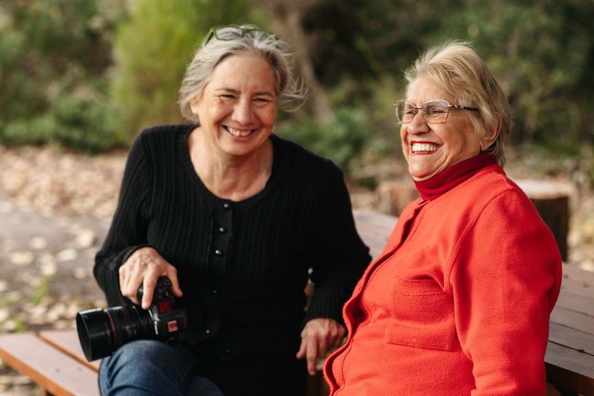 A close-up shot of two women together. Both are smiling. One wears red clothes. The other wears black and holds a camera.