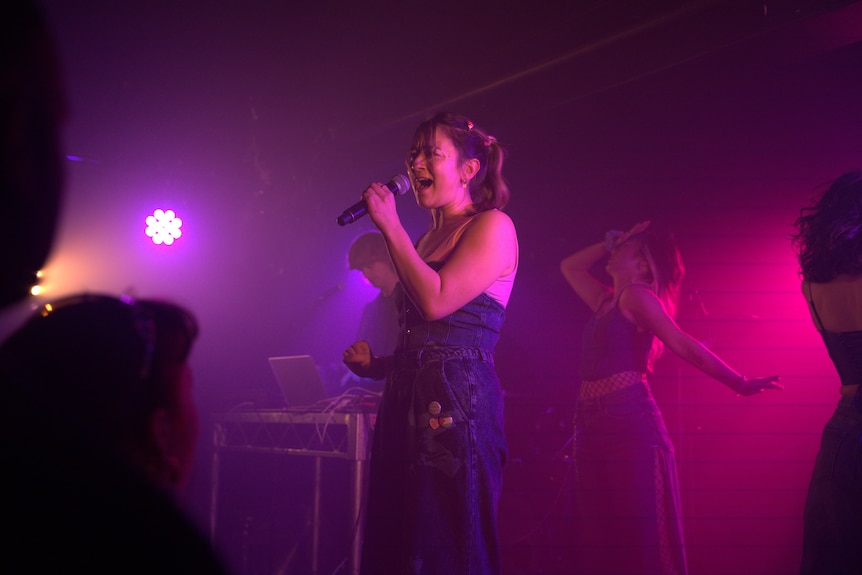 Emi Doi stands on stage with two dancers and holds a microphone.