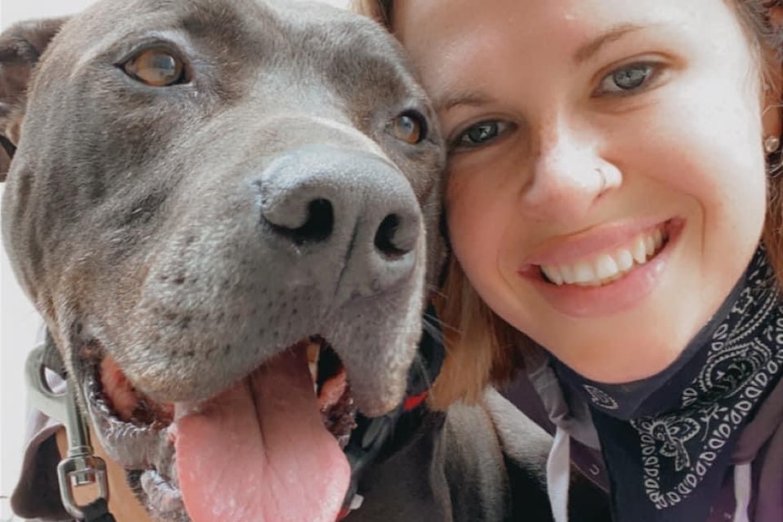 Kara, a young woman smiles as she poses for a selfie with a large grey dog with it's tongue hanging out.