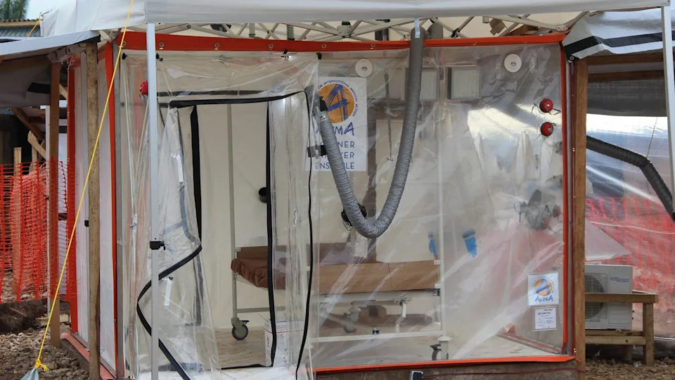 A plastic, transparent cube-like structure with a zip-up door that is open. There is an empty bed inside and no patient