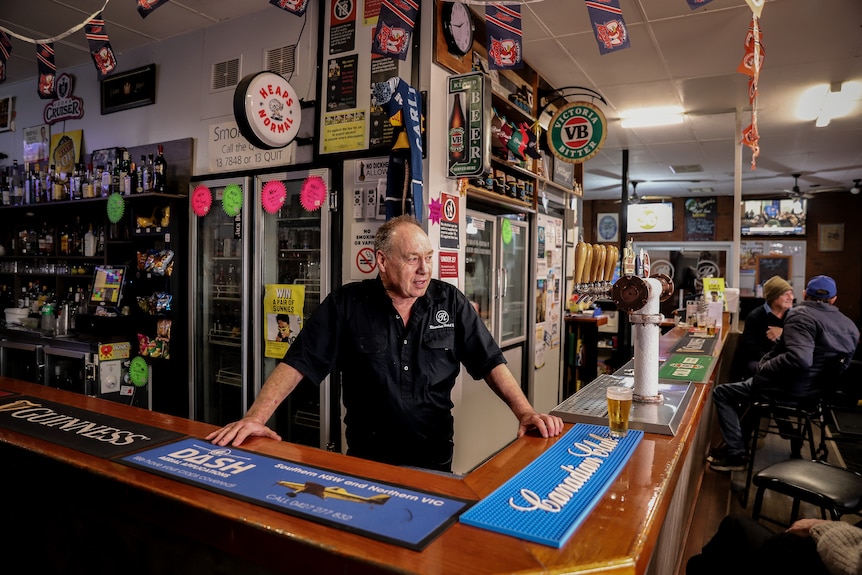 A man in a black short sleeved shirt stands behind a bar with fridges and signage behind him.