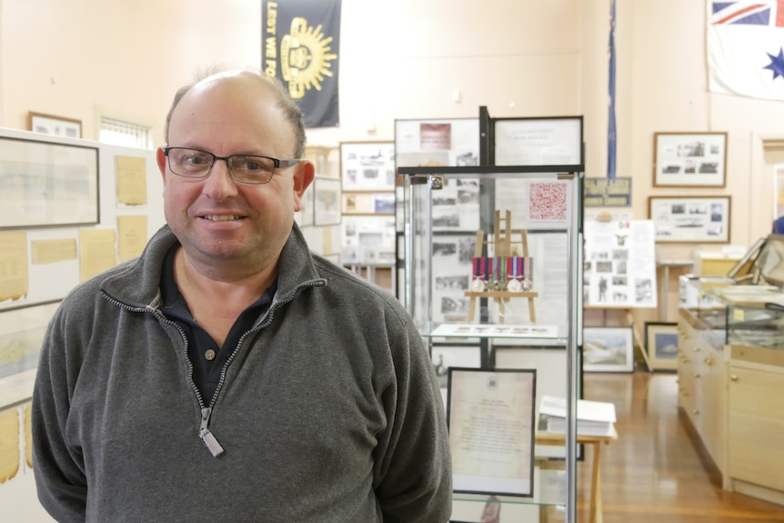 A man with glasses and black jumper stands in front of a glass case with medals and certificates in it