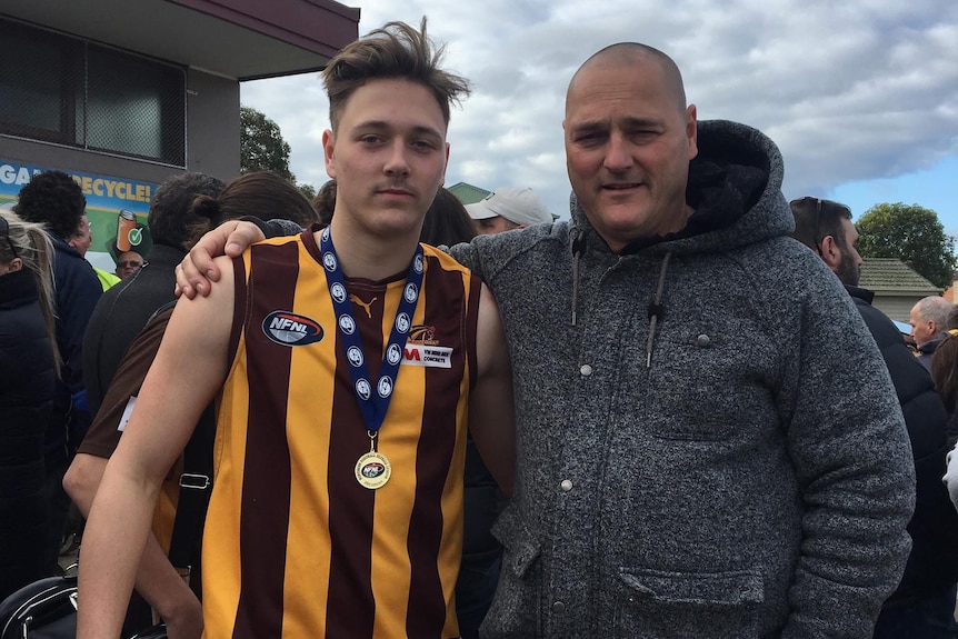 A man with closely shaved hair has his arm around a man in a yellow and brown striped footy top with a medal around his neck.