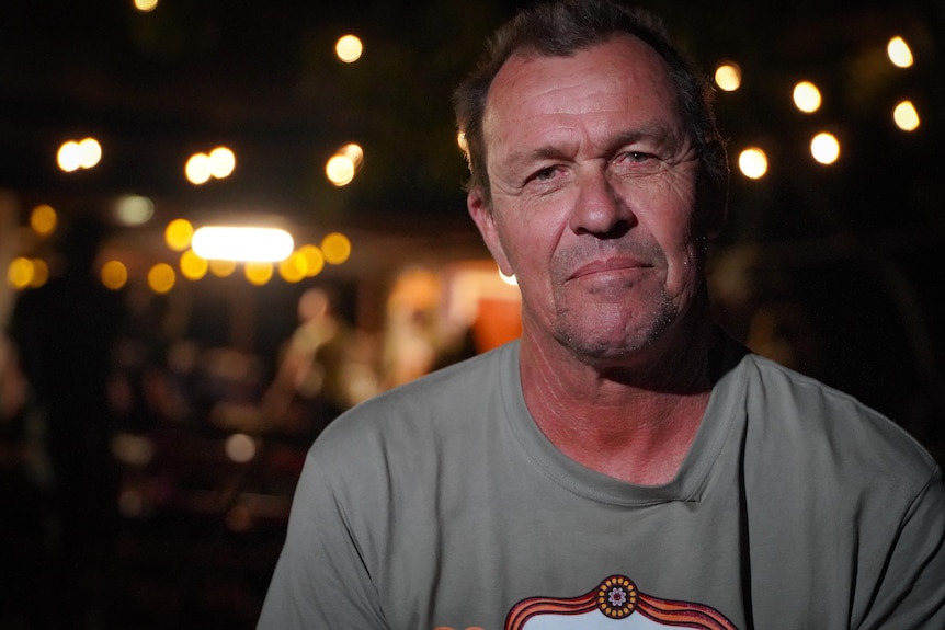 A man looks at the camera with yellow globes lighting up the night behind him.