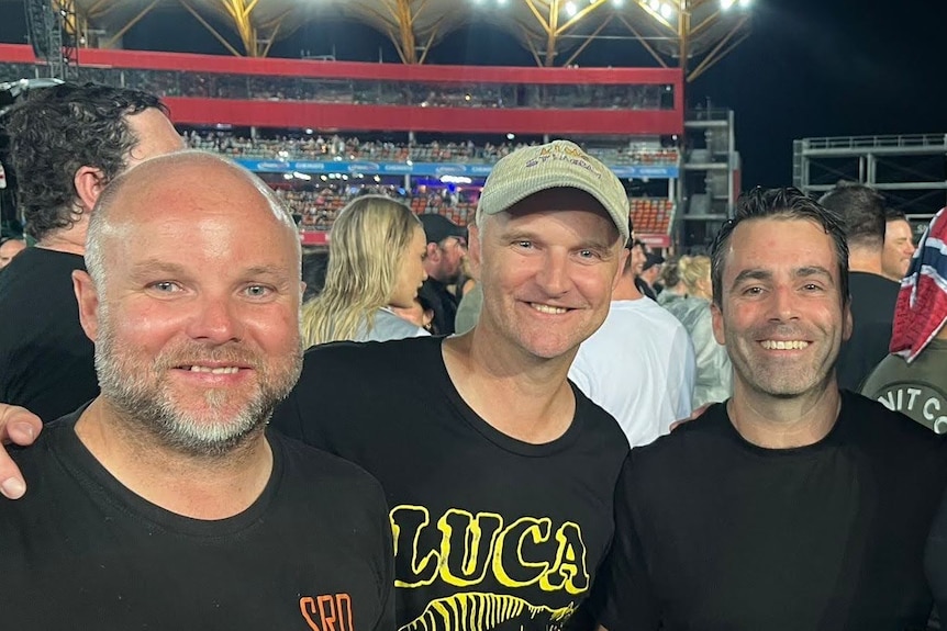 Three smiling men stand among a ccrowd of concert-goers in a stadium.