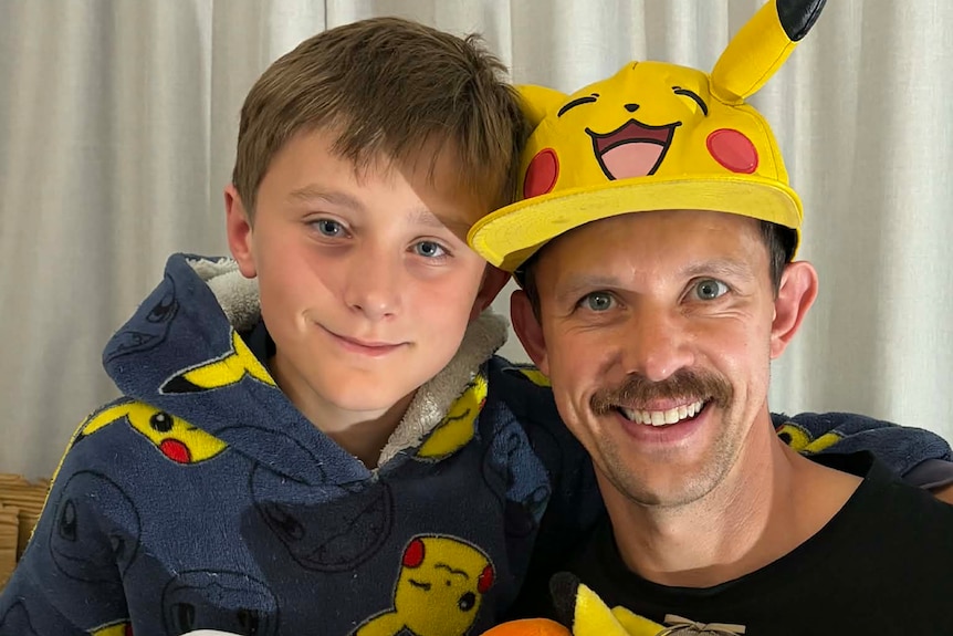 A man wearing a Pikachu hat from the Pokemon series smiles with his son.