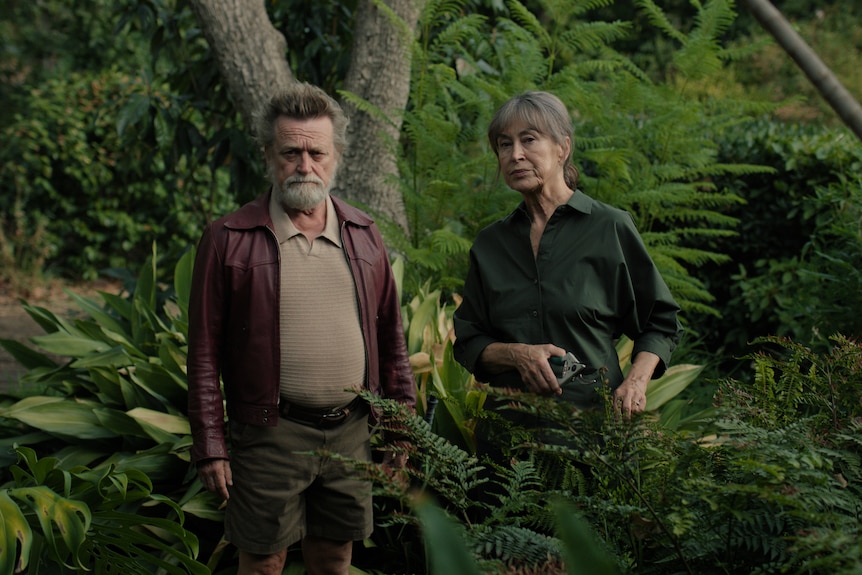 A film still of Michael Hurst, 68, bearded, hair raised, and Linda Cropper, 68, wary, standing in a well-maintained backyard.