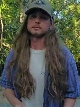 A man with long hair and a green cap looks at the camera with trees behind him