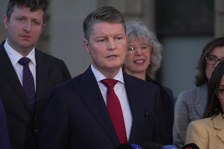 A man with dark wavy hair in a black blazer, white collared shirt and red tie stands in front of a group of people.