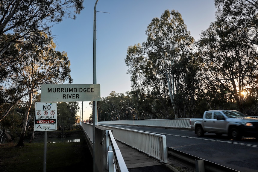 A sign next to a bridge reads 'MURRUMBIDGEE RIVER' a white utility vehicle passes on the opposite side of the road.