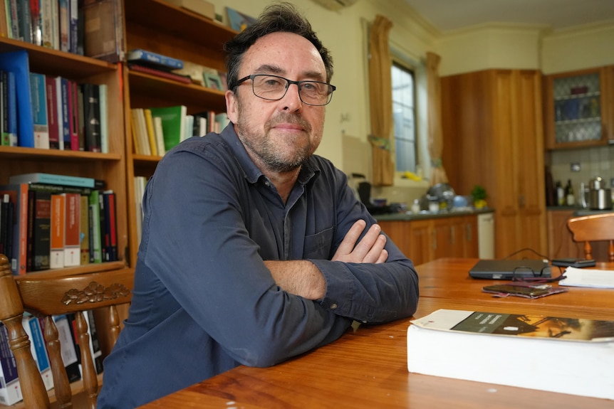 A man wearing glasses and a long sleeved shirt sitting at a desk with a bookshelf behind him.