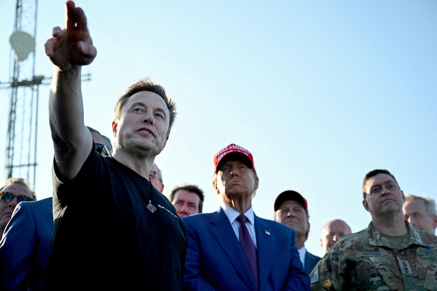 Elon Musk points at something in the distance while Donald Trump, wearing a blue suit and red MAGA cap, watches on.