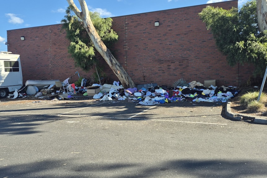 Rubbish and household items piled against a brick wall, in a carpark.