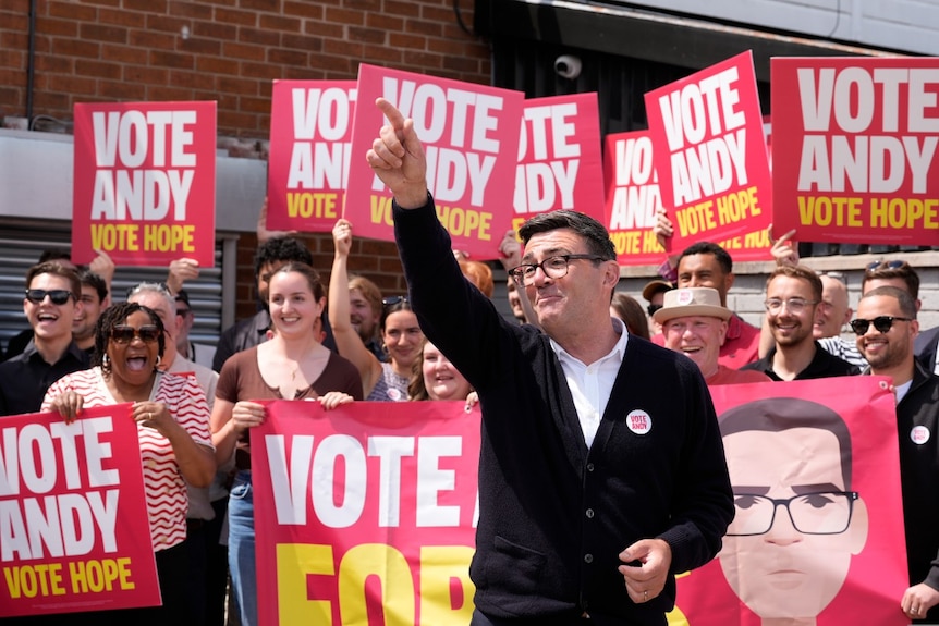 A man smiling and pointing, in front of a large crowd of people holding signs in support of him.