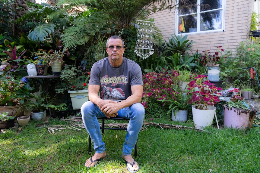 A man with short dark grey hair wearing a mauve tshirt, blue jeans and black thongs sits on a chair in his backyard.
