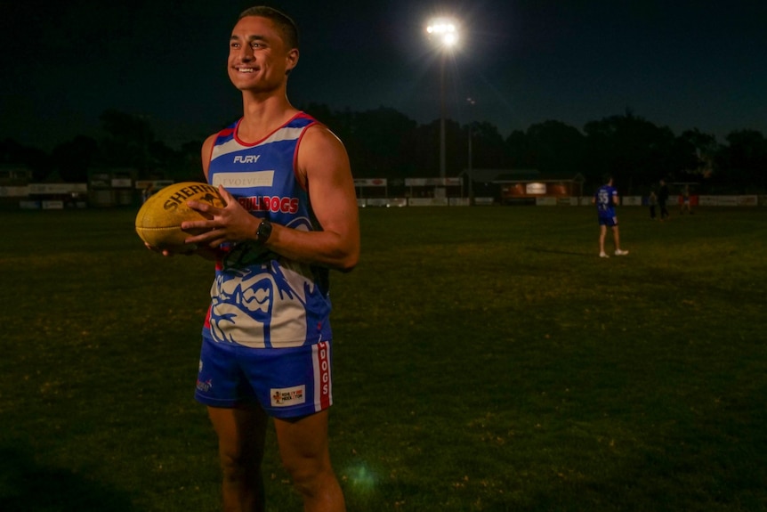 A young man holds a football on a field at night.
