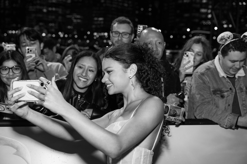 A black and white photo of Catherine Lagaʻaia taking a selfie with fan, other fans behind her